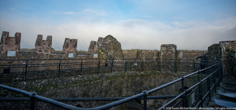 Black visitor railings wind around weathered stone parapets and crenellations on the rooftop of Blarney Castle.