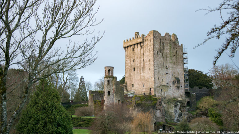 Tall stone keep of Blarney Castle with crenellated roofline and adjoining ruins, framed by trees and shrubs.