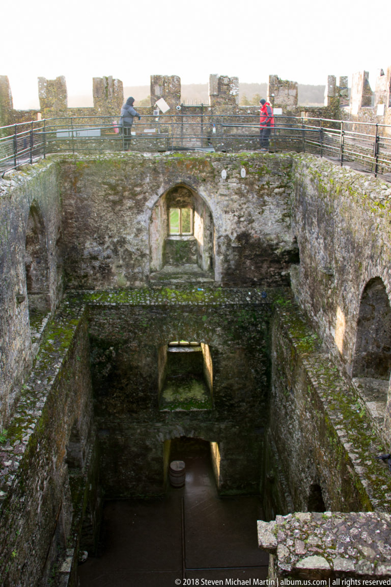 Roofless stone chamber inside Blarney Castle with arched openings, deep walls, and visitors on railings above.