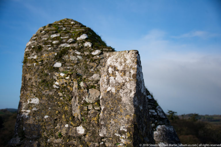 Rounded weathered stone wall fragment topped with grass and moss against blue sky.