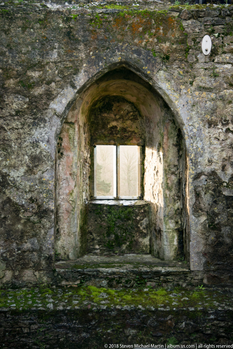 Arched stone wall recess containing a small rectangular window, with worn surfaces, deep sill, and moss staining.