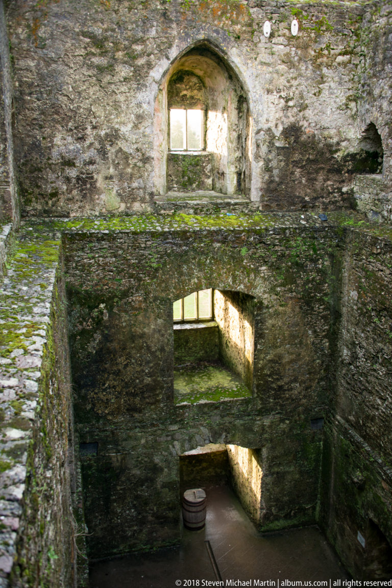 Roofless castle interior with stacked stone openings, deep walls, sunlight, mossy ledges, and a barrel-like object below.