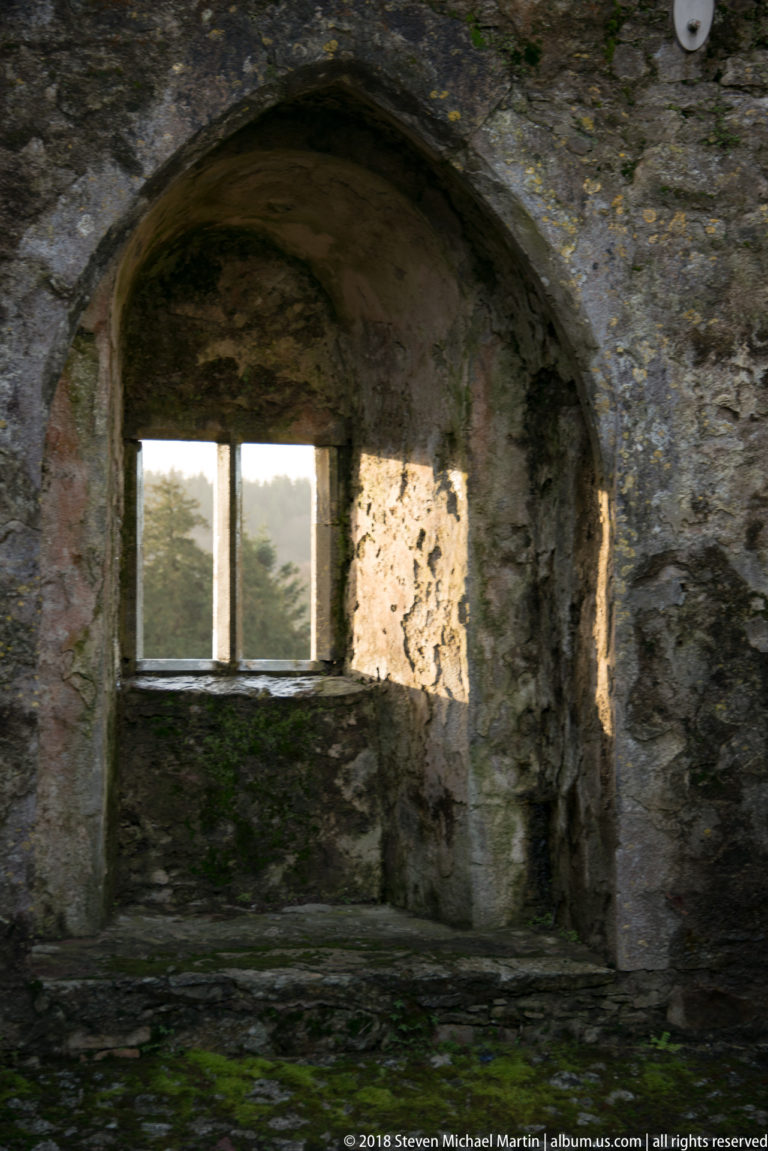 Sunlit arched wall recess with a small rectangular window set deep within rough stone masonry.