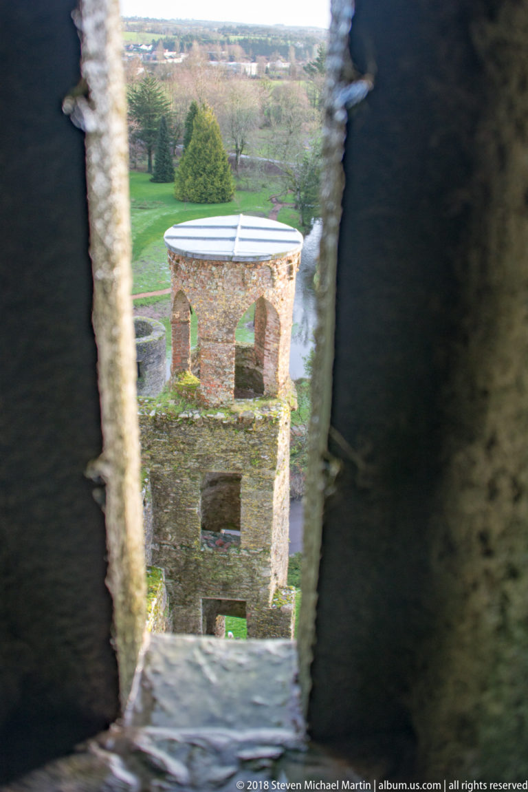 Small ruined stone tower on the grounds viewed through a narrow shadowed opening in Blarney Castle.