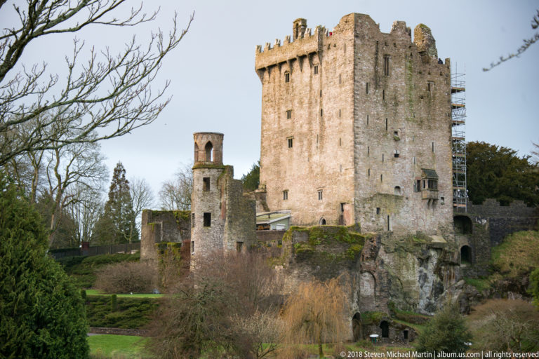 Weathered Blarney Castle keep with crenellations, narrow windows, adjoining tower, and lower ruined walls above garden vegetation.