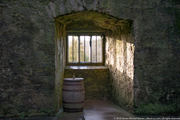 Deep stone window recess with iron bars and a wooden barrel beneath the opening.