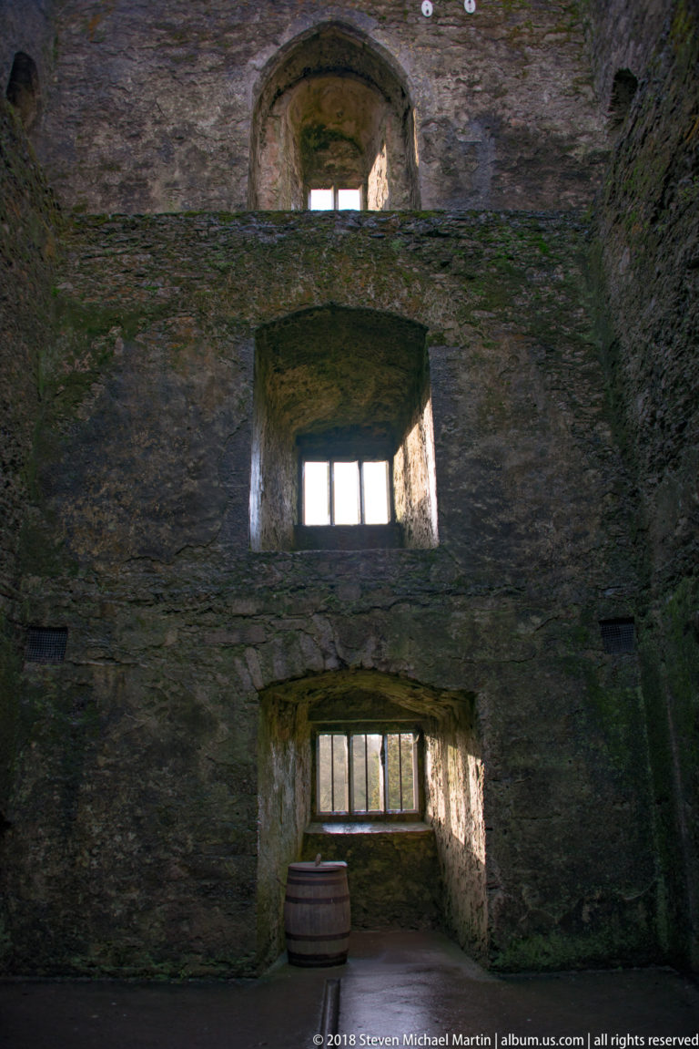 Tall rough-stone wall with three deep window openings arranged vertically and a barrel at floor level.