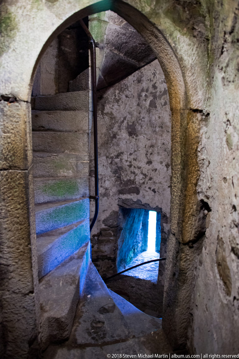 Narrow stone spiral staircase curving upward through thick castle masonry beside a simple handrail.