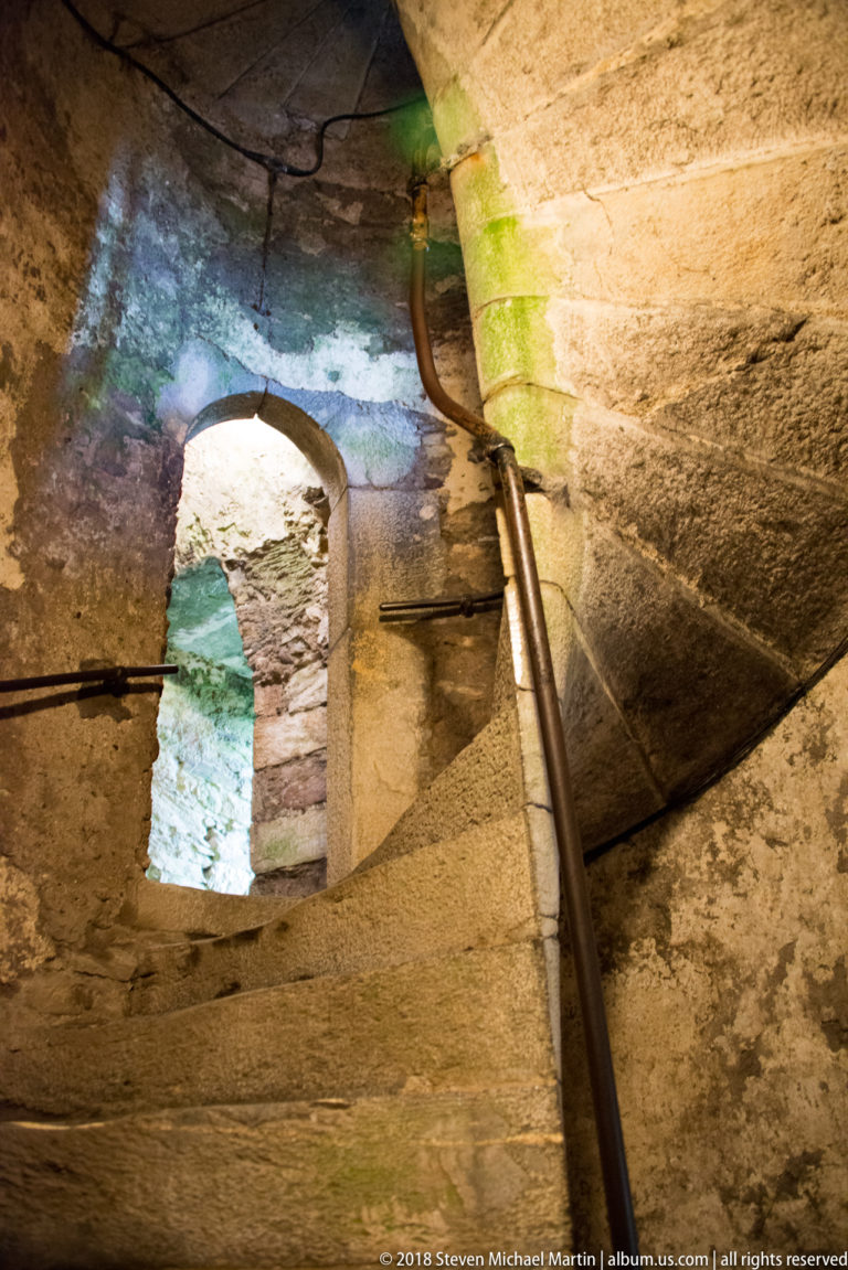 Close view of worn spiral-stair treads beside a narrow stone window opening and metal handrail.