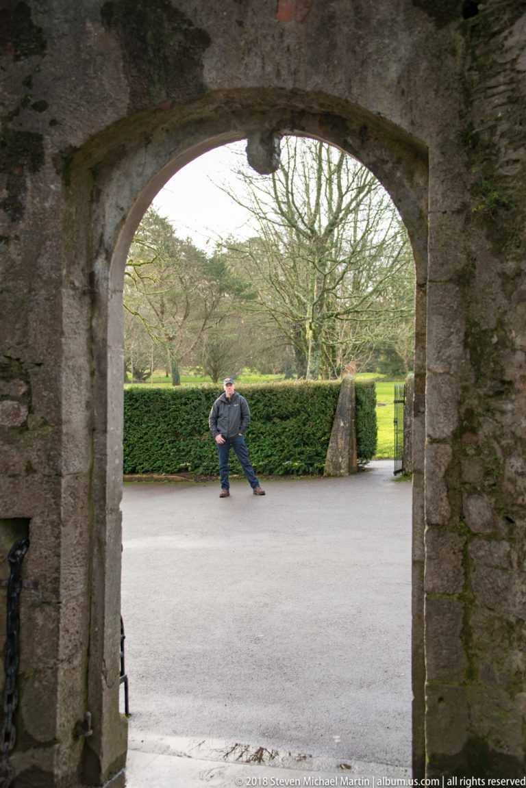 Arched stone doorway opening from the castle toward hedges, a path, and a standing visitor outside.