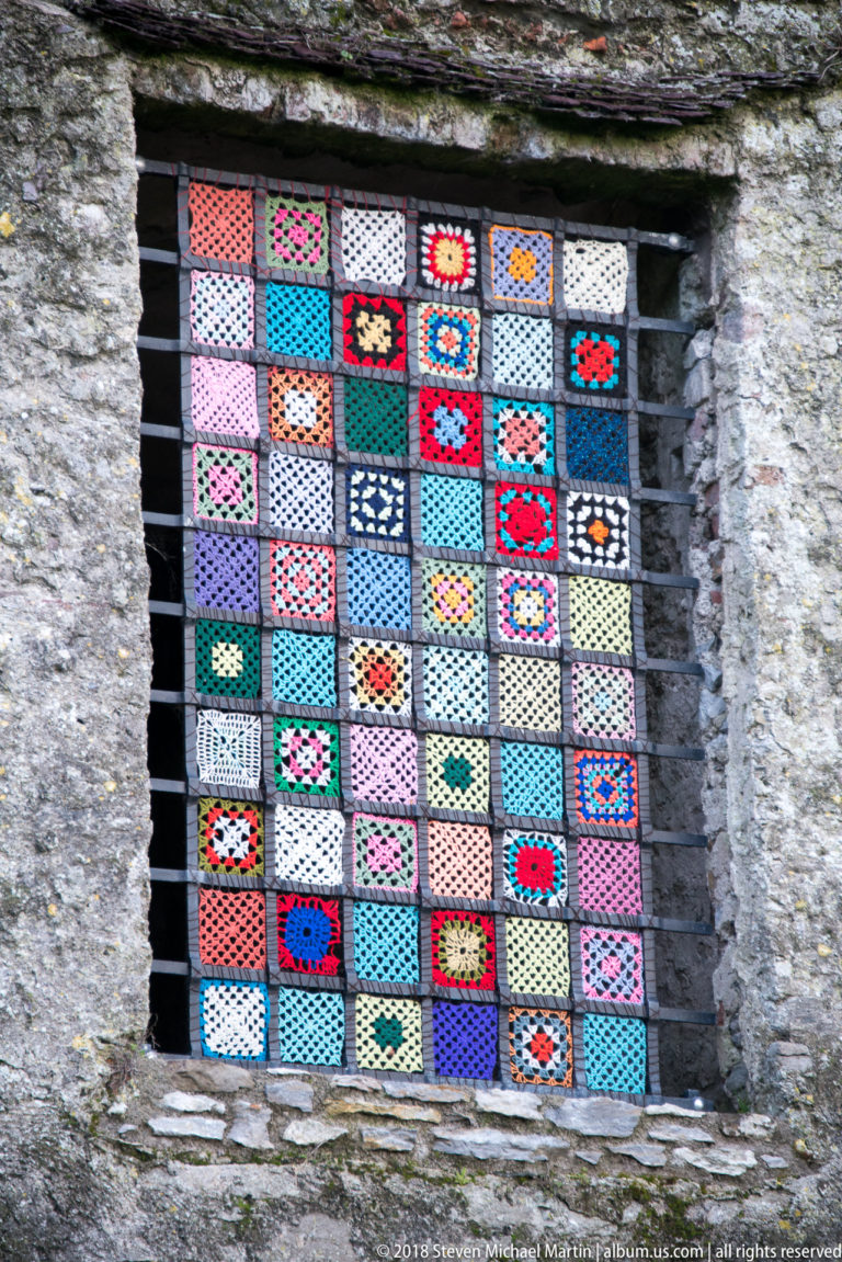 Colorful crocheted square panels arranged behind a black metal grid in a rough stone window opening.