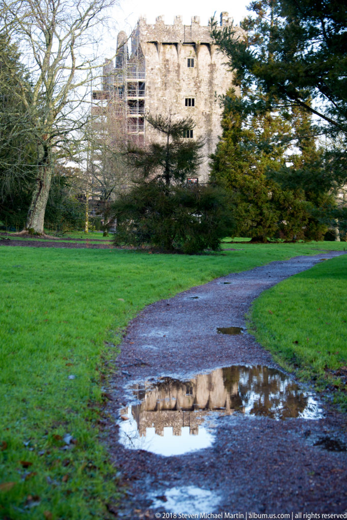 Blarney Castle partly visible through trees and reflected in a puddle on a curving path.