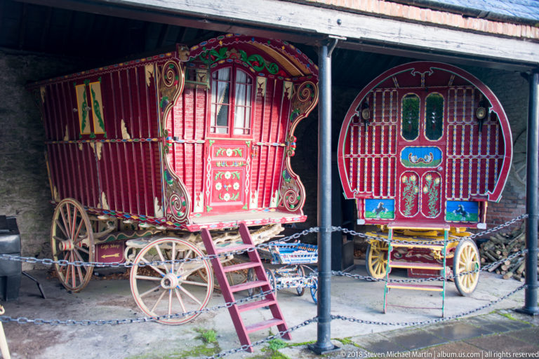 Two ornate red horse-drawn caravans with painted panels, windows, spoked wheels, and entry steps under a shelter.
