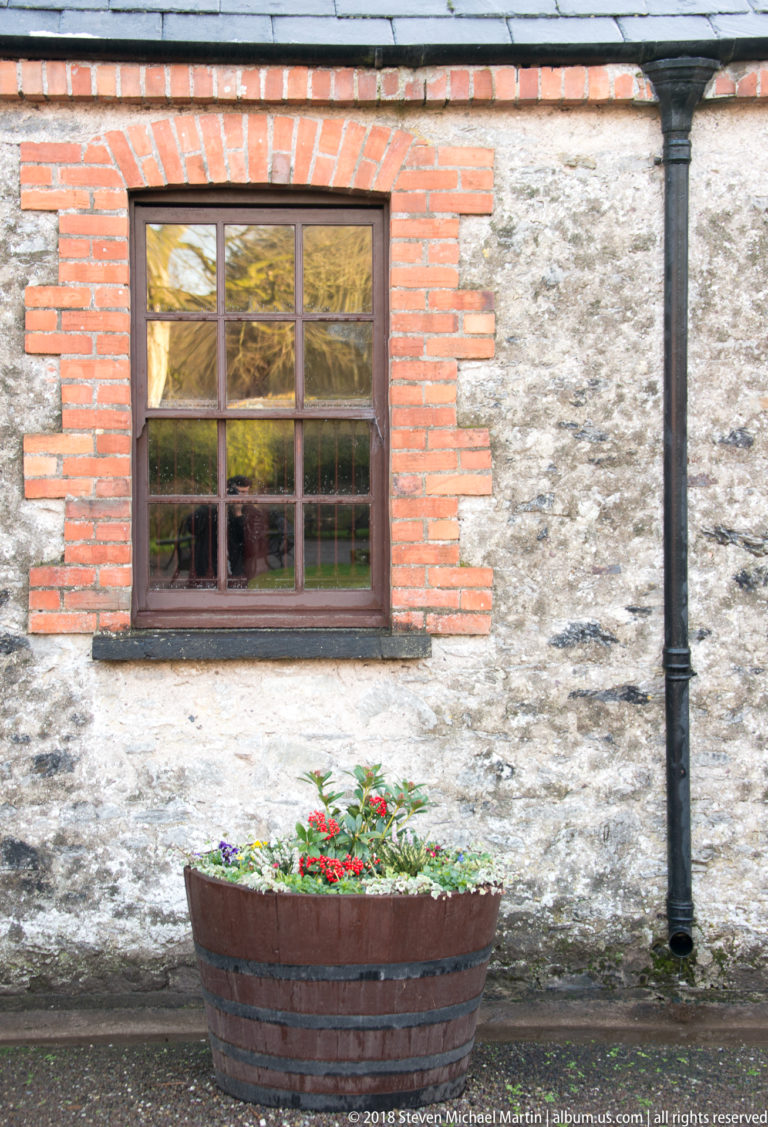 Wooden window framed in red brick above a barrel planter with flowers against a rough stone wall.