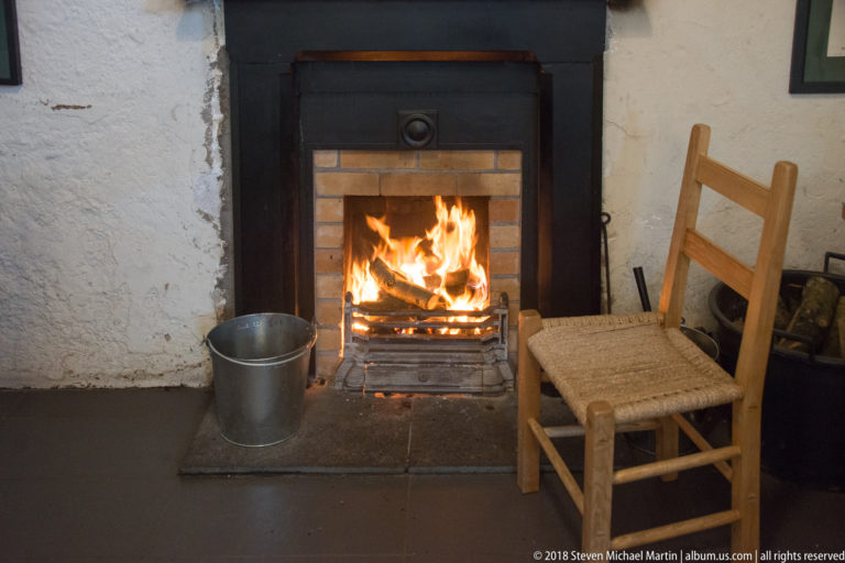 Lit fireplace in a dark surround beside a wooden chair and metal bucket on a tiled hearth.