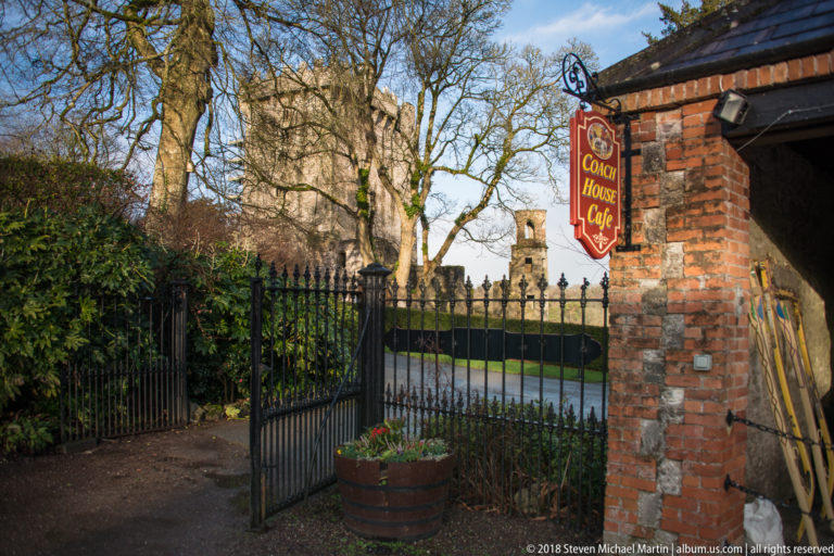 Coach House Café sign beside brick pillar and iron gate, with Blarney Castle visible in the distance.
