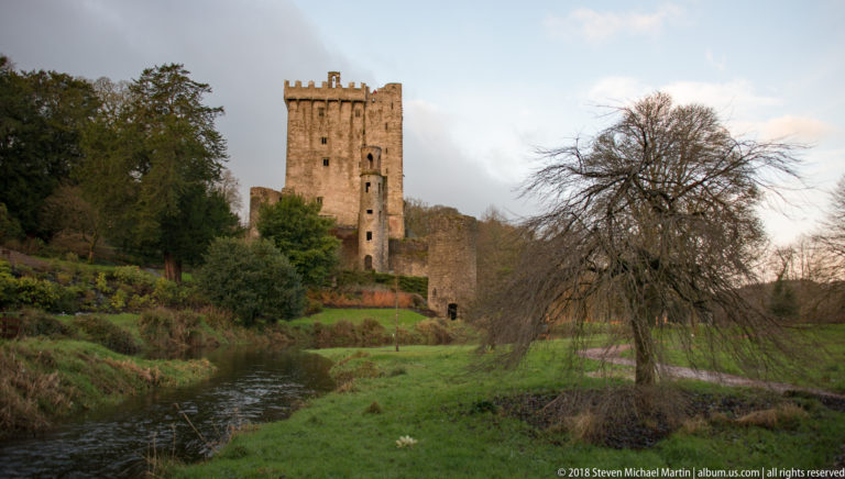 Blarney Castle keep across a dark stream, with green banks, evergreens, and a leafless tree under pale evening light.
