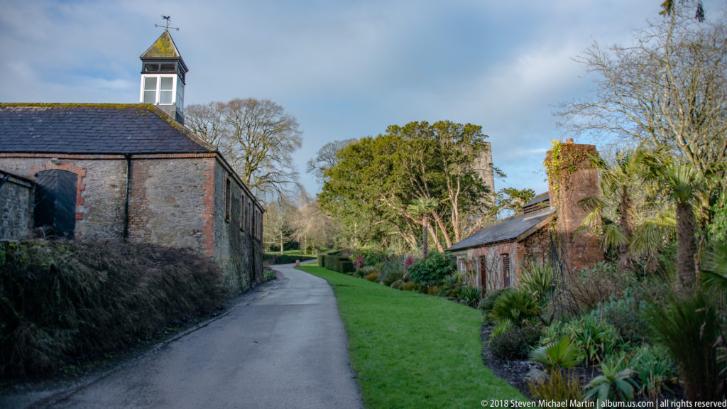 Narrow paved lane between stone estate buildings, bordered by lawn, shrubs, and mature trees.
