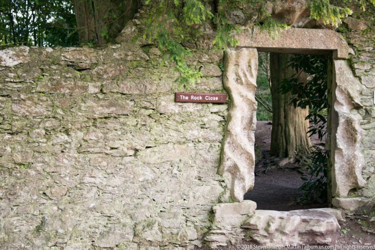 Rough stone wall with a small doorway labeled The Rock Close, opening onto a shaded garden path.