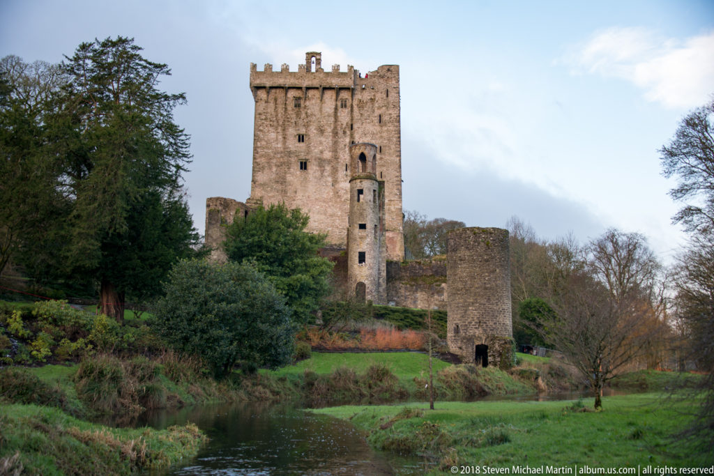 Blarney Castle keep with lower stone outworks above a stream and green garden banks.