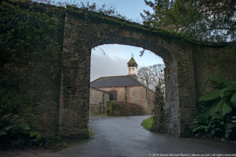 Large rough-stone archway over a paved road, framing a small gabled building with a roof lantern beyond.