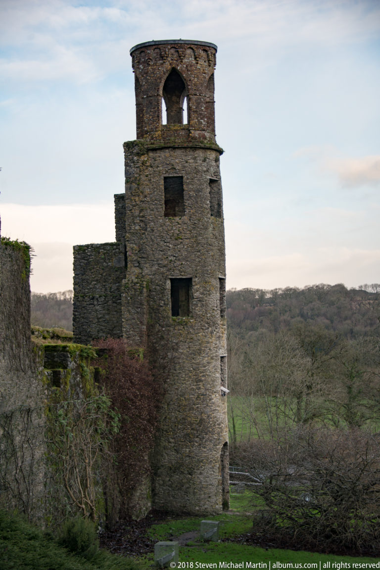 Narrow ruined stone tower with multiple openings and a large upper arch, attached to lower wall fragments.