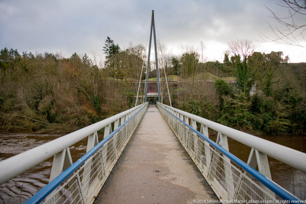Pedestrian suspension bridge crossing the River Boyne between wooded banks under an overcast sky.