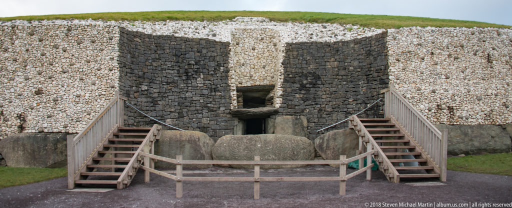Wide frontal view of Newgrange entrance, carved entrance stone, stairs, and contrasting stone façade.