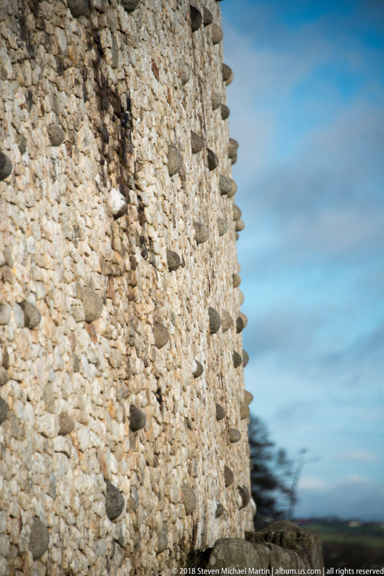 Close oblique view along Newgrange’s pale quartz-and-cobble outer wall against blue sky.
