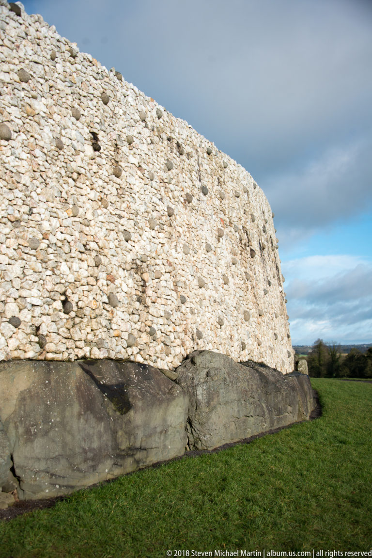 Curving row of massive kerbstones below Newgrange’s pale revetment, with grass beside the mound.
