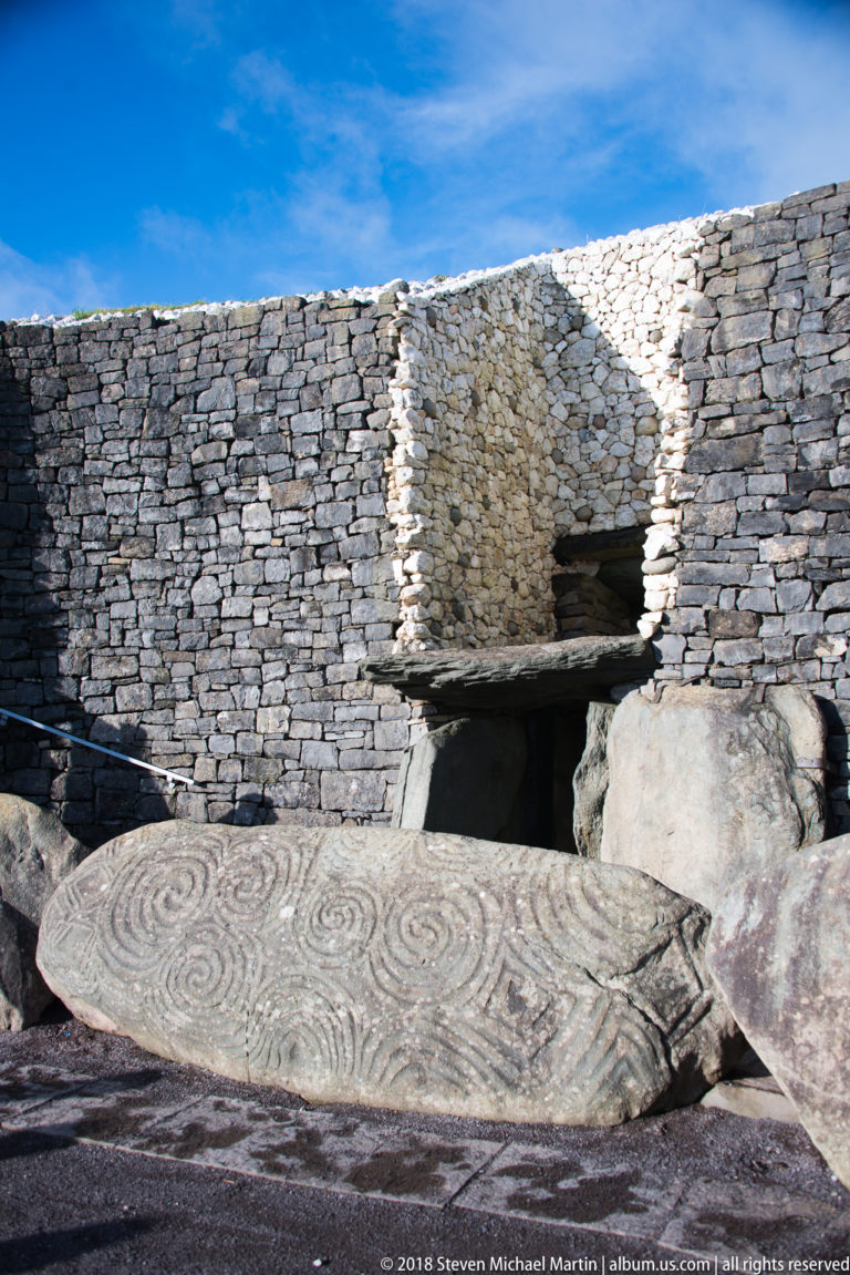 Sunlit Newgrange entrance with carved entrance stone, dark passage, and contrasting stone façade.