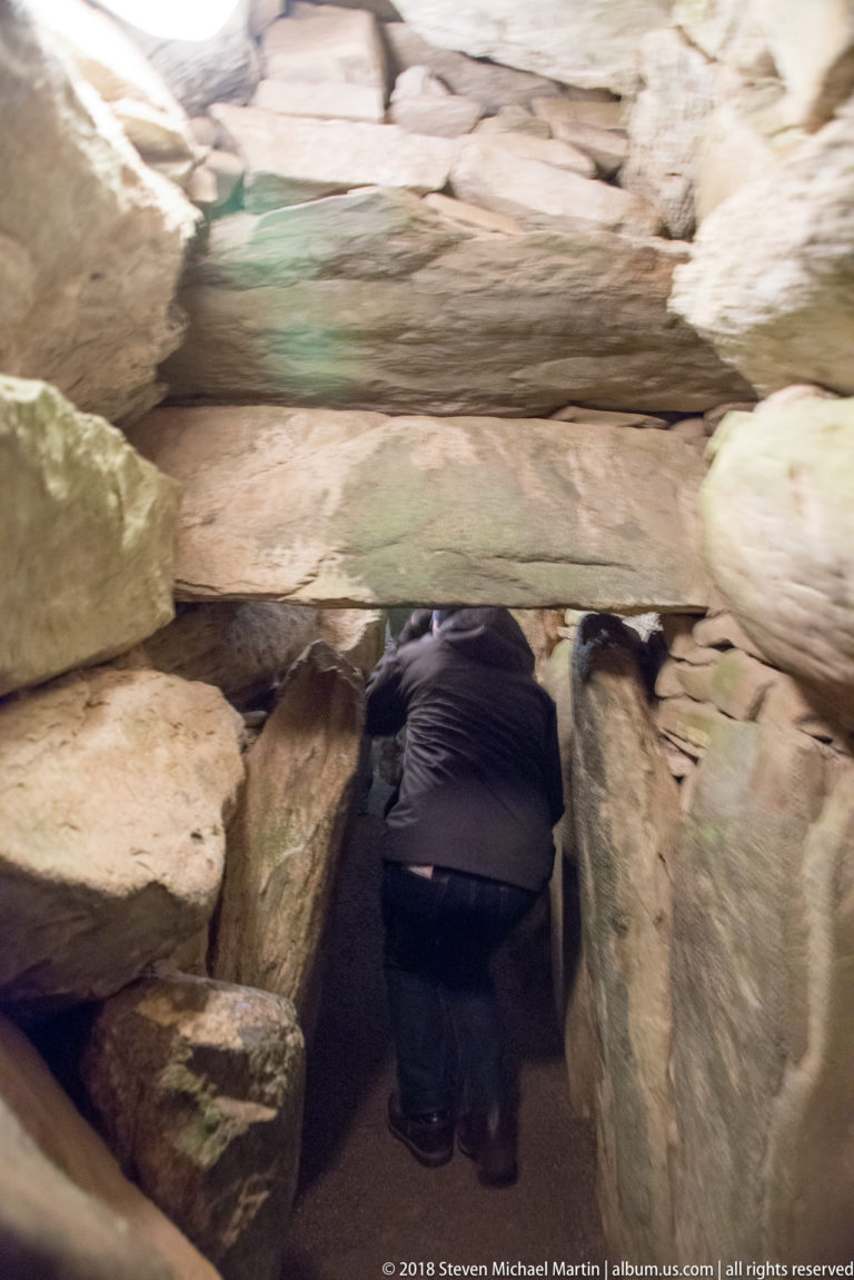Visitor bending through a low, narrow megalithic passage lined with large upright stones at Newgrange.