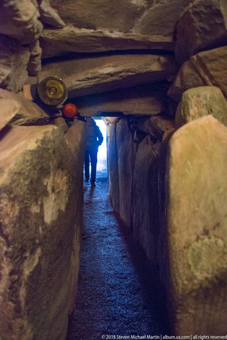 View outward through Newgrange’s narrow stone passage toward daylight and a small visitor silhouette.