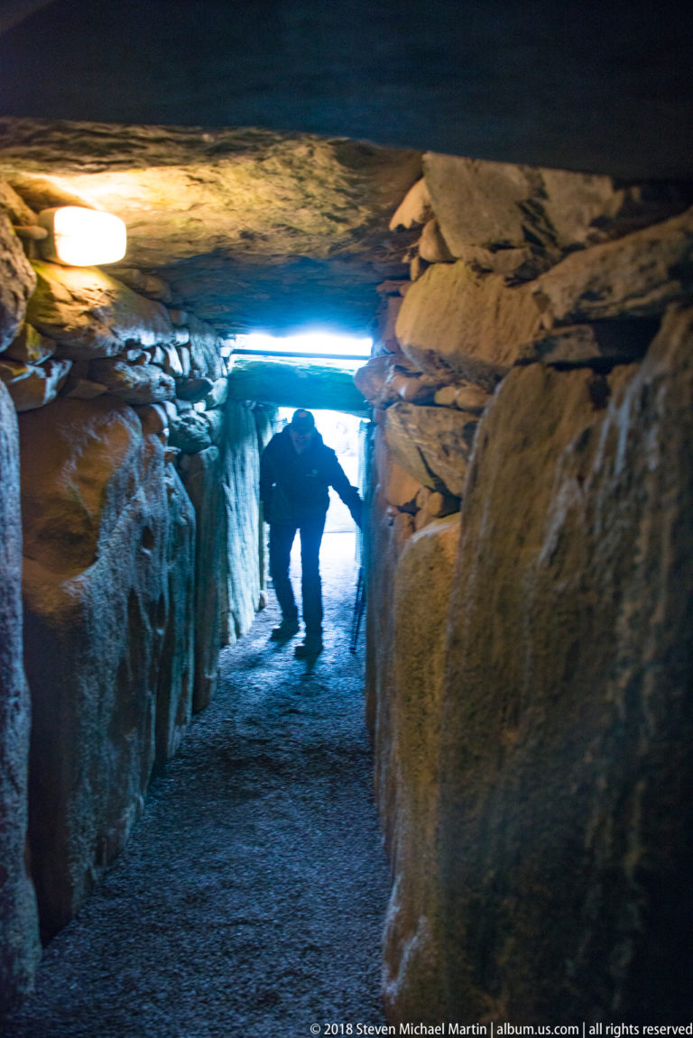 Dim stone passage inside Newgrange with artificial lights and a visitor walking toward the bright entrance.