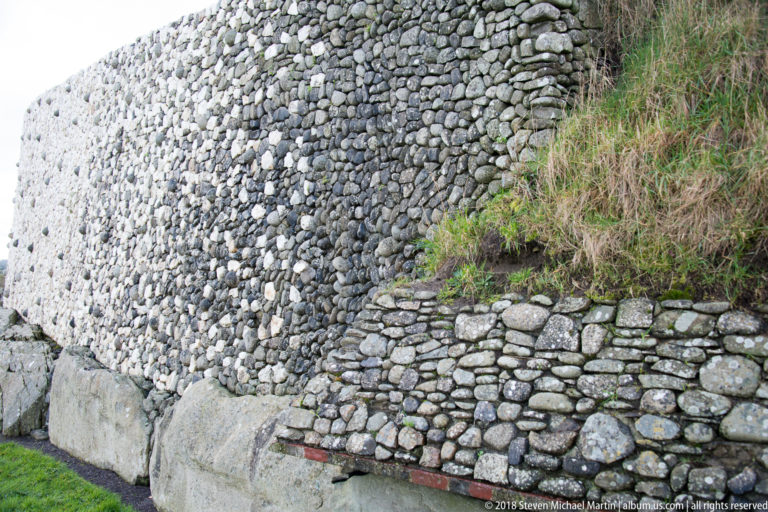 Side detail where Newgrange’s pale pebble façade meets lower stonework and the grassy mound.