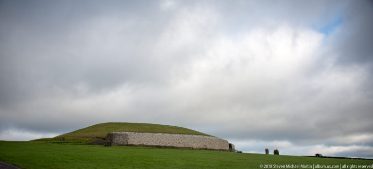 Grass-covered Newgrange mound with a white-quartz façade beneath a dramatic cloudy sky.