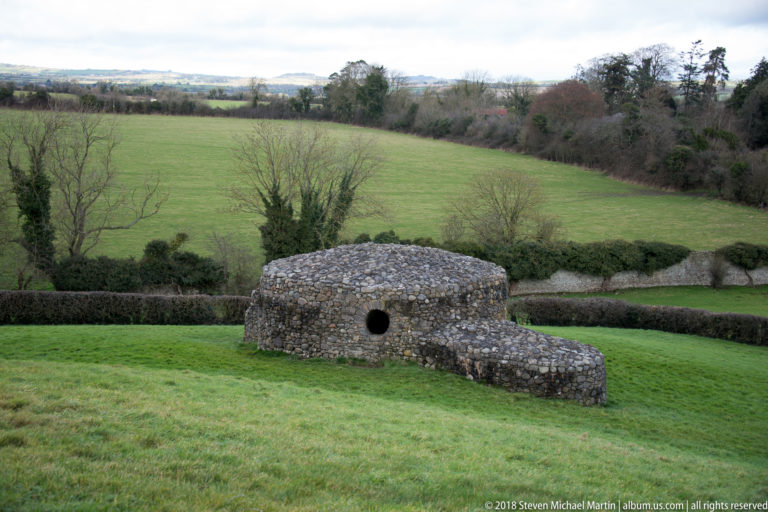 Low circular dry-stone structure with a small dark opening on grass above green fields and hedgerows.