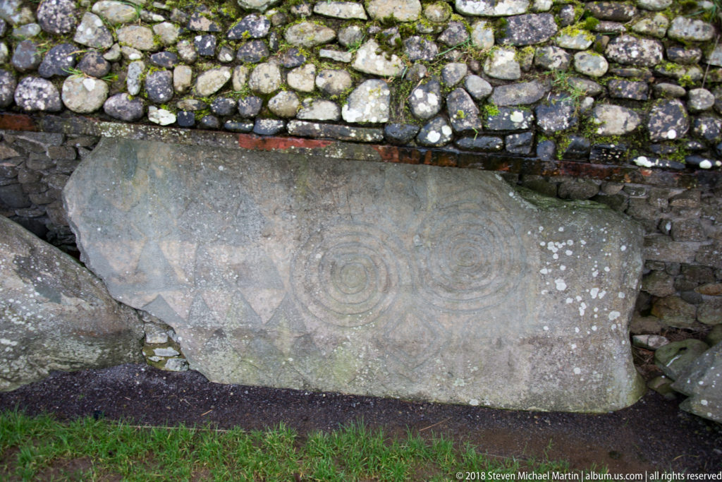 Large weathered kerbstone beneath a low stone wall, carved with concentric spirals and angular geometric motifs.