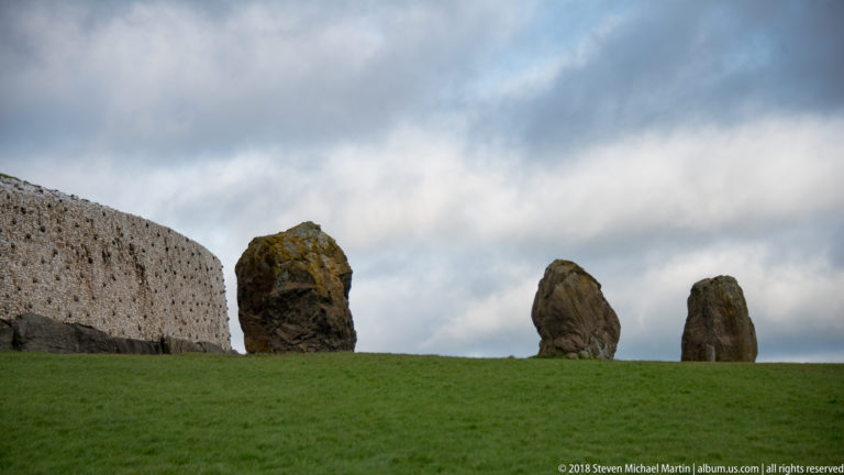 Three upright standing stones on grass beside the pale stone facing of the Newgrange mound under a cloudy sky.