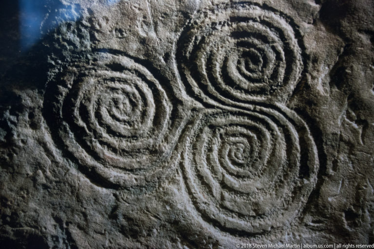 Close view of several deeply carved spiral motifs on a dark, rough-surfaced megalithic stone.