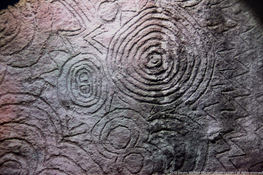 Large concentric spiral surrounded by smaller coils and curving incisions on a rough megalithic stone surface.