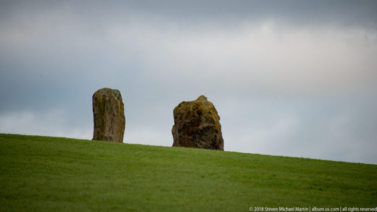 Two irregular standing stones rising from a grassy slope beneath a gray sky.