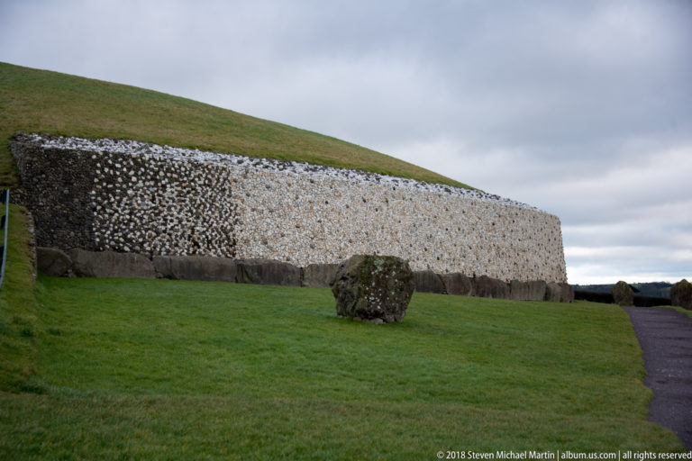 Oblique view of Newgrange’s quartz-and-cobble façade with a large rough stone in the foreground.