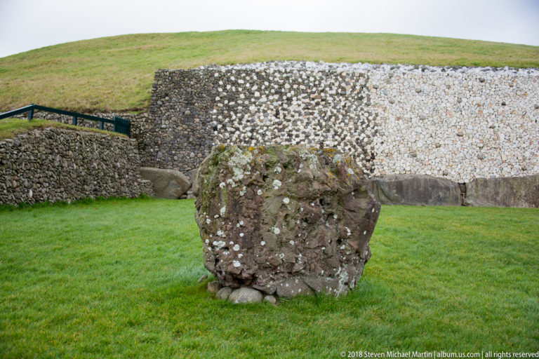 Lichen-spotted boulder on grass in front of Newgrange’s pale quartz-and-cobble façade.