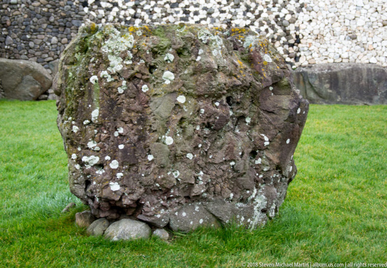 Close view of a rough lichen-covered boulder with Newgrange’s pebble façade behind it.
