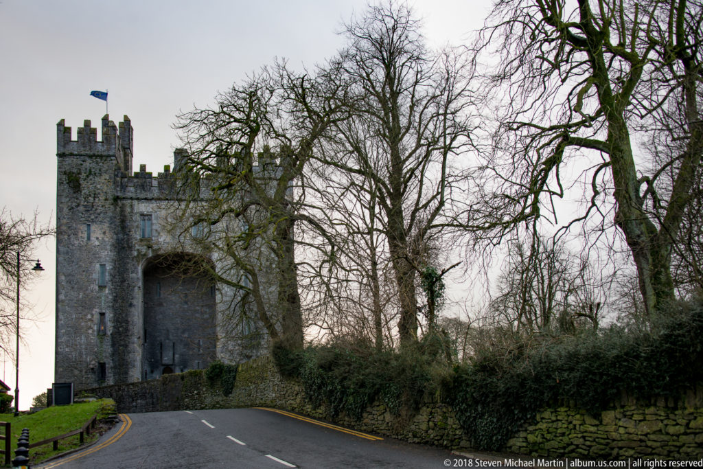 Crenellated stone gate tower with a large central arch spanning a curving road among bare trees.
