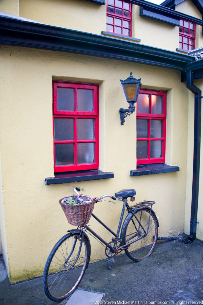Bicycle with wicker basket leaning below red-framed windows on a yellow pub wall with a lantern.
