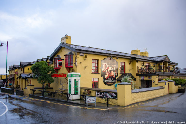 Wide view of yellow Durty Nelly's pub with red windows and signage beside a wet street in Bunratty.