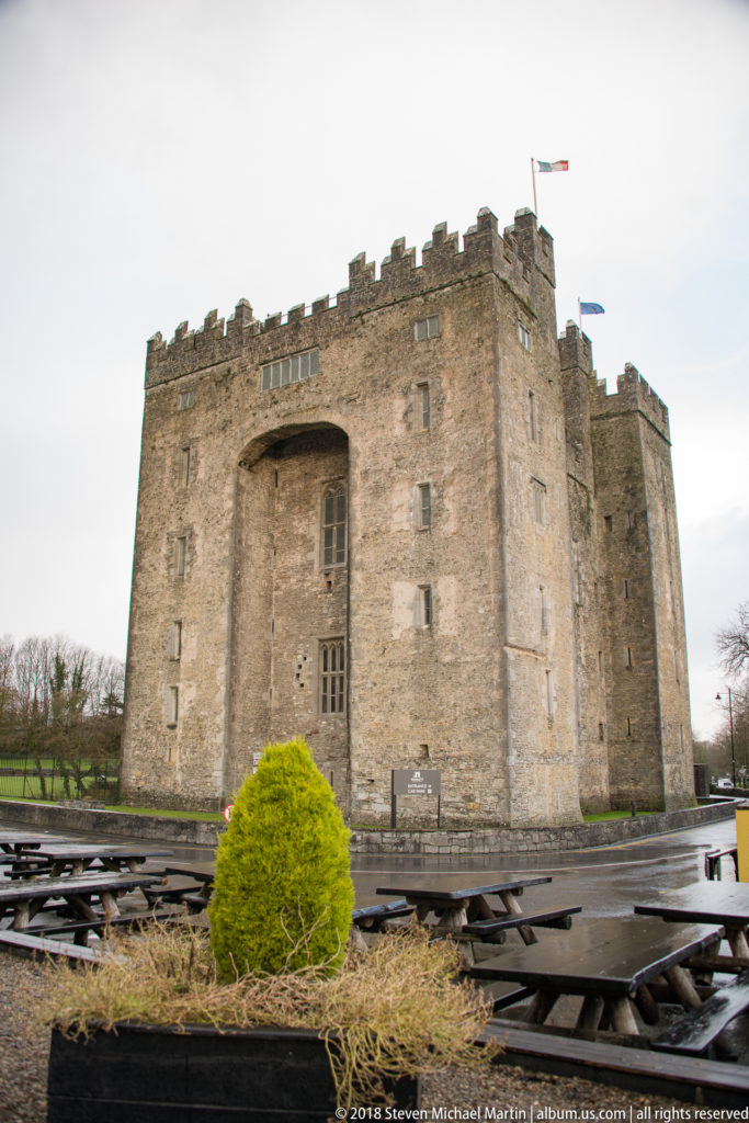 Massive crenellated stone Bunratty Castle rises behind rows of wet outdoor benches under an overcast sky.