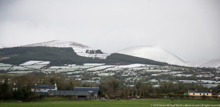 Snow-covered hills rise beyond fields, hedgerows, farm buildings, and scattered houses near Cahir.